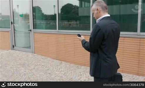 Happy businessman walking and speaking on cell phone near modern office building. Steadicam shot