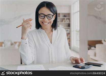Happy businesswoman in glasses counting profit on calculator, analyzing benefits, enjoying financial success. Female employer smiling working with financial documents, counts company budget.. Happy businesswoman counts profit on calculator, analyzes company budget, enjoying financial success