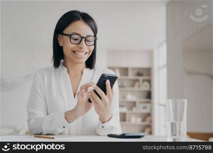 Happy businesswoman in glasses uses mobile apps for business, holding smartphone, sitting in office. Smiling female reading message with good news, chatting online in workplace, looking at phone.. Happy businesswoman in glasses uses mobile apps for business, holding smartphone, sitting in office