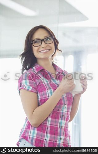 Happy businesswoman looking away while holding coffee mug in creative office