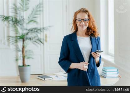 Happy businesswoman reads incoming notification, holds smartphone device, checks email box, has wavy red long hair, dressed in formal clothes, poses over office interior, has confident look at camera