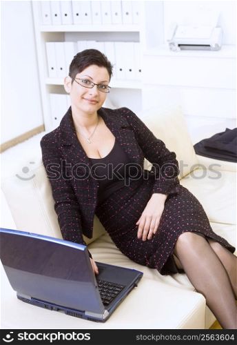 Happy businesswoman sitting on sofa at office and working on laptop computer, smiling.