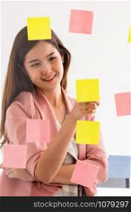Happy businesswoman thinking creative ideas with sticky notes on glass wall at the office. Work planning and education concept