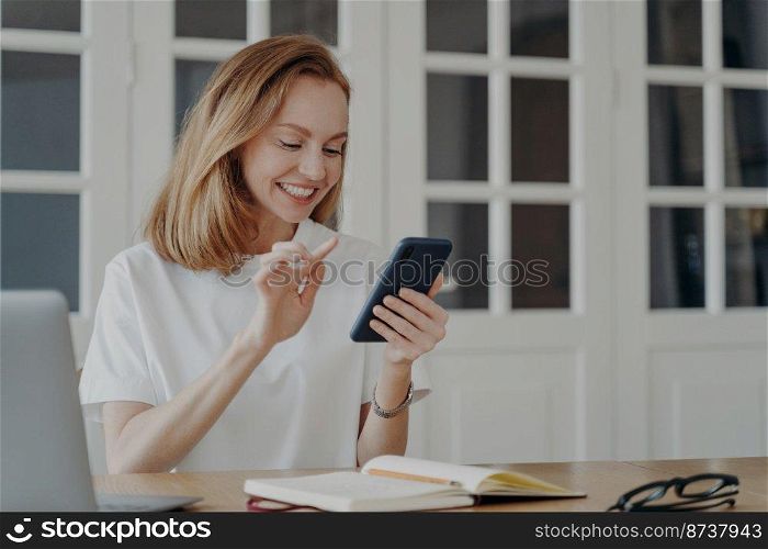 Happy businesswoman uses smartphone in home office, sitting at desk. Smiling female having fun with cell phone, using business or entertainment mobile apps in the workplace, browsing the Internet.. Smiling female holding smartphone using mobile apps browsing the Internet in the workplace
