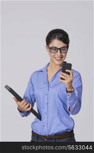 Happy businesswoman using mobile phone while holding laptop against gray background