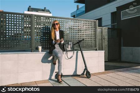 Happy businesswoman using mobile sitting on a wall with her scooter next. Happy businesswoman using mobile with her scooter next