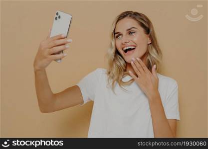 Happy carefree woman dressed casually with smartphone making selfie in studio against nude background, taking picture of herself for social media, looking at camera on mobile phone with cute smile. Happy carefree woman dressed casually with smartphone making selfie in studio against nude background
