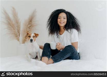 Happy carefree woman with Afro hairstyle, wears t shirt nad jeans, feels glad, holds mug of coffee, laughs sincerely, jack russell terrier poses near host. Cheerful teenage girl with favourite pet