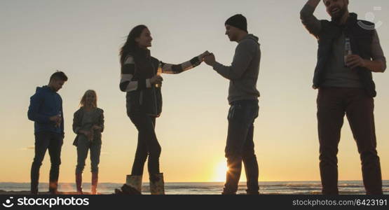 Happy Carefree Young Friends Having Fun And Drinking Beer By Bonefire On The Beach As The Sun Begins To Set