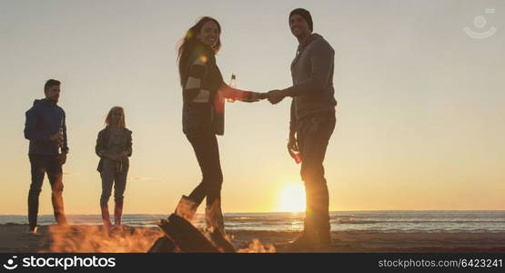 Happy Carefree Young Friends Having Fun And Drinking Beer By Bonefire On The Beach As The Sun Begins To Set