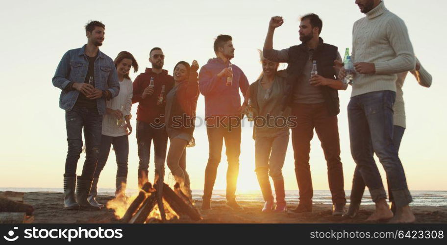 Happy Carefree Young Friends Having Fun And Drinking Beer By Bonefire On The Beach As The Sun Begins To Set