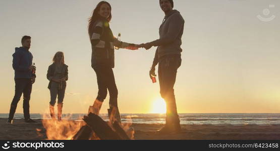 Happy Carefree Young Friends Having Fun And Drinking Beer By Bonefire On The Beach As The Sun Begins To Set