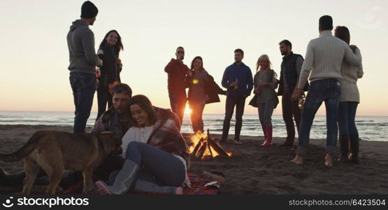 Happy Carefree Young Friends Having Fun And Drinking Beer By Bonefire On The Beach As The Sun Begins To Set