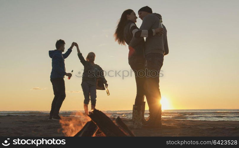 Happy Carefree Young Friends Having Fun And Drinking Beer By Bonefire On The Beach As The Sun Begins To Set