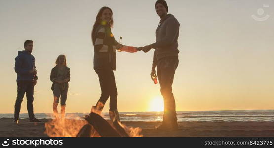 Happy Carefree Young Friends Having Fun And Drinking Beer By Bonefire On The Beach As The Sun Begins To Set