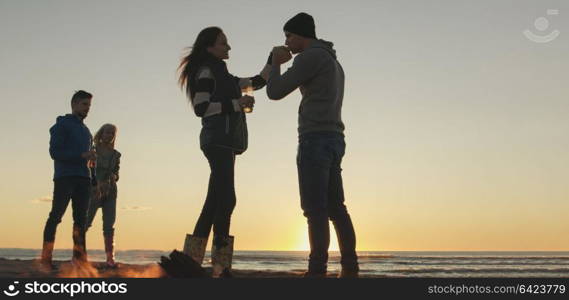 Happy Carefree Young Friends Having Fun And Drinking Beer By Bonefire On The Beach As The Sun Begins To Set