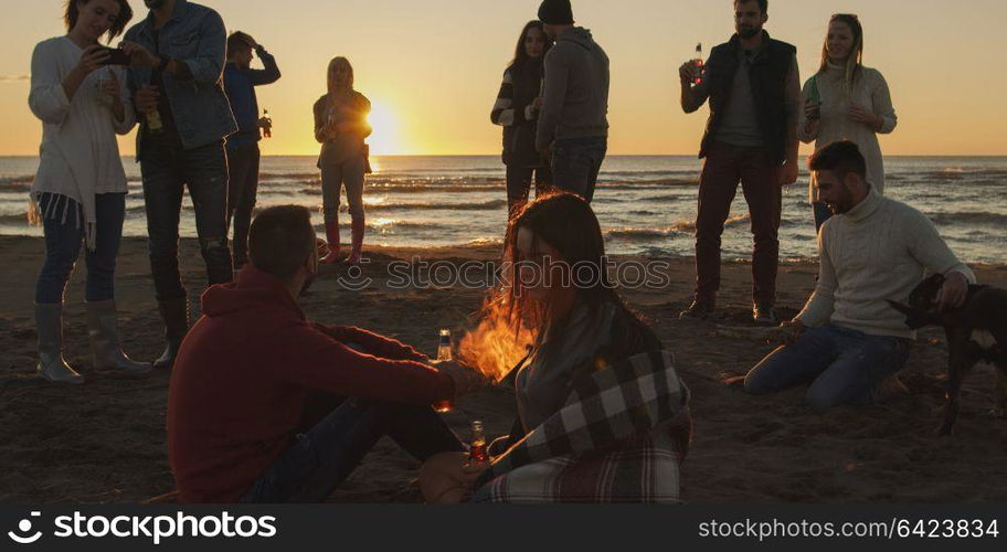 Happy Carefree Young Friends Having Fun And Drinking Beer By Bonefire On The Beach As The Sun Begins To Set