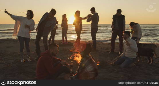 Happy Carefree Young Friends Having Fun And Drinking Beer By Bonefire On The Beach As The Sun Begins To Set