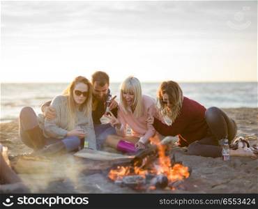 Happy Carefree Young Friends Having Fun And Drinking Beer By Bonefire On The Beach As The Sun Begins To Set. Friends having fun at beach on autumn day