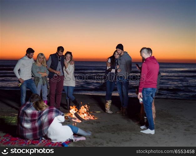 Happy Carefree Young Friends Having Fun And Drinking Beer By Bonefire On The Beach As The Sun Begins To Set. Friends having fun at beach on autumn day