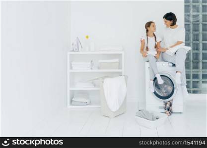 Happy caring woman embraces her little daughter, sit on washing machine, have rest after washing, surrounded with basket and dirty clothes, have pleasant conversation with each other. Housework