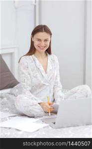 Happy casual beautiful woman working on a laptop sitting on the bed in the house. woman checking social apps and working. Communication and technology concept. Woman in quarantine for Coronavirus.. Happy casual beautiful woman working on a laptop sitting on the bed in the house. woman checking social apps and working. Communication and technology concept. Woman in quarantine for Coronavirus