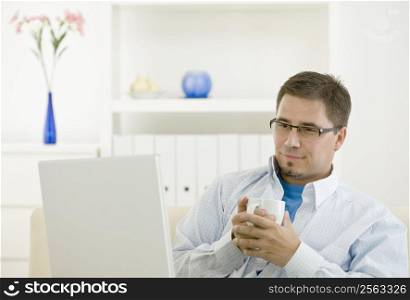 Happy casual man using laptop computer at home drinking coffee.