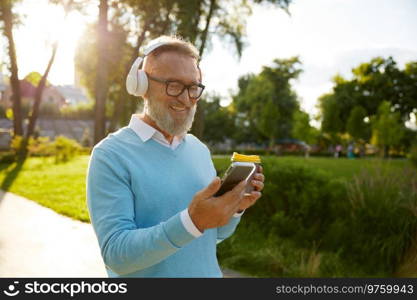 Happy casual senior man wearing headset having video conference or watching funny movie on mobile drinking coffee on walk in public park. Happy senior man having video conference on mobile drinking coffee on walk