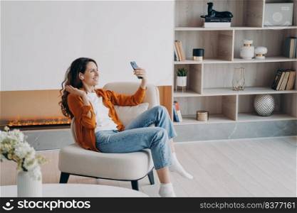 Happy caucasian girl has video phone call at home in weekend morning. Young woman is using airpods. Wireless earphones. Technology using, leisure and communication concept. Beautiful modern interior.. Happy caucasian girl has video phone call at home in weekend morning. Young woman is using airpods.