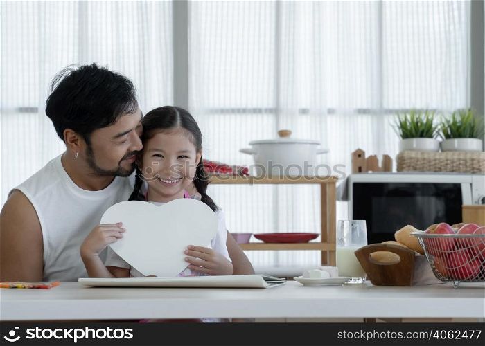Happy Caucasian little daughter smiling holding heart shaped card and giving to her Asian young father after finished drawing and making gift for dad on fathers day at home kitchen with love