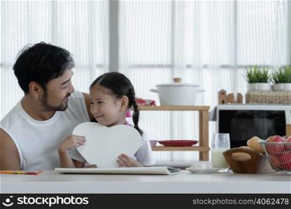Happy Caucasian little daughter smiling holding heart shaped card and giving to her Asian young father after finished drawing and making gift for dad on fathers day at home kitchen with love