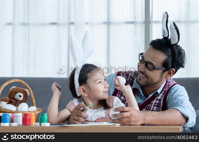 Happy Caucasian little girl daughter decorating by coloring Easter eggs with young Asian father in bunny ears headbands in living room at home. Mixed race family prepare for Easter holiday at home