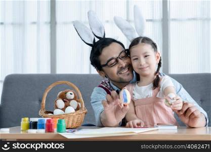 Happy Caucasian little girl daughter holding showing decorated Easter eggs with young Asian father in bunny ears headbands in living room at home. Mixed race family prepare for Easter holiday at home