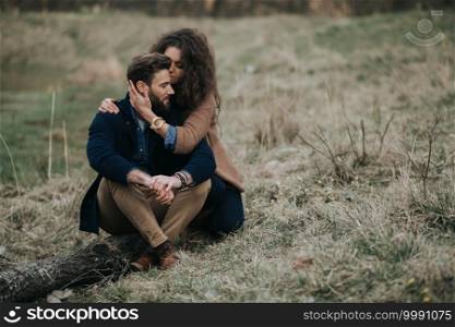 Happy caucasian lovers are sitting on the shore of the lake. Young couple is hugging on autumn day outdoors. A bearded man and curly woman in love. Valentine’s Day. Concept of love and family. Happy caucasian lovers are sitting on the shore of the lake. Young couple is hugging on autumn day outdoors. A bearded man and curly woman in love. Valentine’s Day. Concept of love and family.