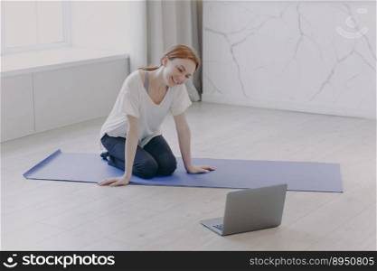 Happy caucasian woman has distance personal training in front of camera. Young pretty sportswoman practicing yoga on mat on floor having video call. Concept of e-learning and home classes.. Happy caucasian woman has distance personal training in front of camera on yoga mat on floor.