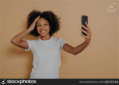 Happy charming dark skinned female 20s in white tshirt making photo selfie on modern smartphone and smiling cheerfuly, isolated over beige wall. Positive young african woman taking photo on cellphone. Happy charming dark skinned female making photo selfie on modern smartphone against beige wall