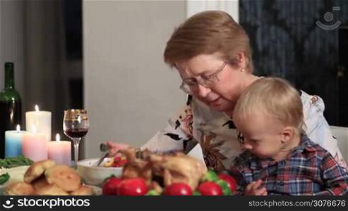 Happy charming senior woman feeding her grandson on Thanksgiving. Loving grandmother sitting at festive table with her cute toddler grandson on her kness and feeding child with healthy food.