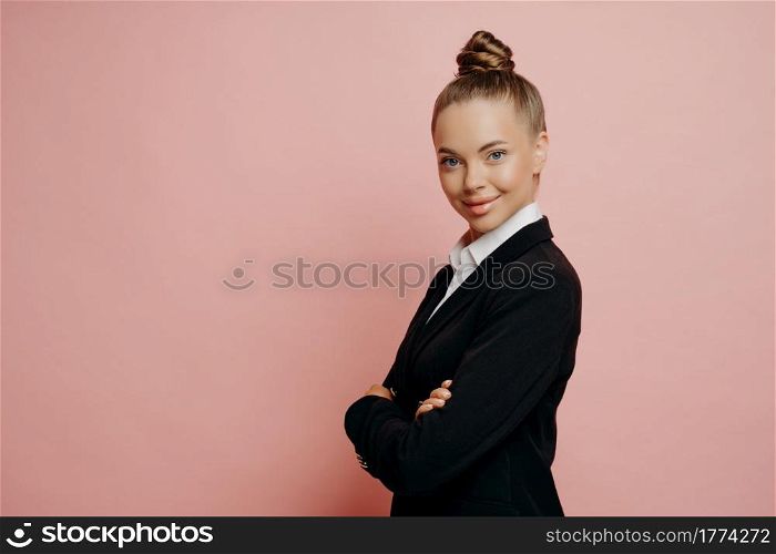 Happy charming woman in suit, professional female business owner in classic wear with hair in bun keeping arms crossed, looking ready to take on challenging work, posing isolated over pink background. Professional female boss looking confidently at camera