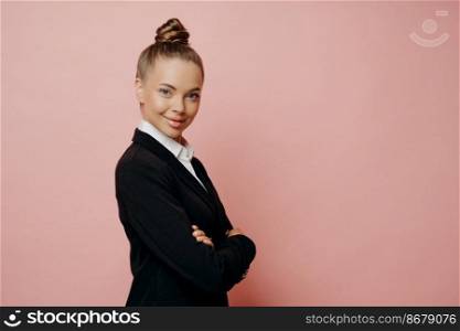 Happy charming woman in suit, professional female business owner in classic wear with hair in bun keeping arms crossed, looking ready to take on challenging work, posing isolated over pink background. Professional female boss looking confidently at camera