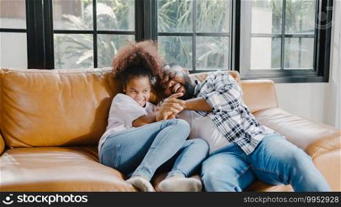 Happy cheerful African American family dad and daughter having fun cuddle play on sofa while birthday at house. Self-isolation, stay at home, social distancing, quarantine for coronavirus prevention.