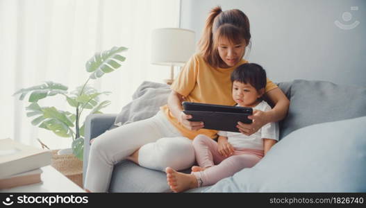 Happy cheerful Asia family mom and cute kid using digital tablet interest cartoon and watch movie having fun relax on couch in living room at house. Spending time together, Quarantine for coronavirus.