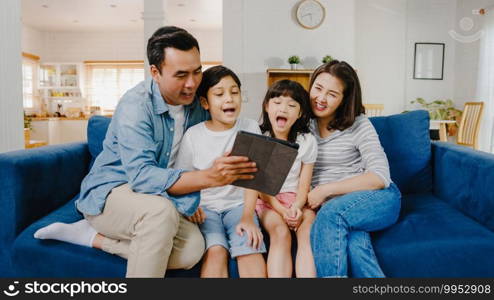 Happy cheerful Asian family dad, mom and kids having fun and using digital tablet video call on sofa at house. Self-isolation, stay at home, social distancing, quarantine for coronavirus prevention.