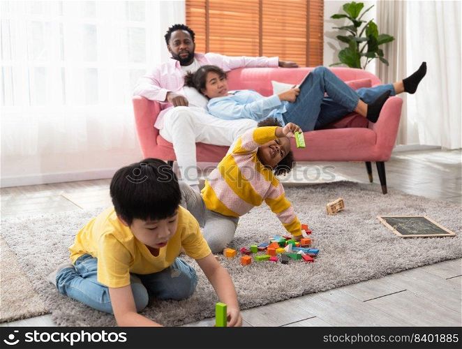 Happy cheerful children playing with colorful wooden building blocks on rug floor and constructing towers in living room. Couple of parents resting on sofa and watching kids playing game with toys at home. Family leisure, games and children development concept