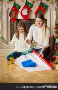 Happy cheerful daughter and mother wrapping sweater in wrapping paper at Christmas eve