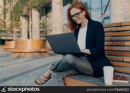 Happy cheerful european ginger woman working remotely on laptop outside, sitting on wooden bench and drinking morning coffee, female in eyeglasses enjoying remote work outdoors, checking e-mail. Happy cheerful european ginger woman working remotely on laptop outside, sitting on wooden bench