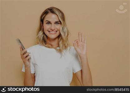 Happy cheerful female in casual tshirt holding smartphone and showing okay gesture, being happy after pleasant conversation or hearing good news, isolated over beige background. Happy cheerful female holding smartphone and showing okay gesture, being happy after pleasant conversation