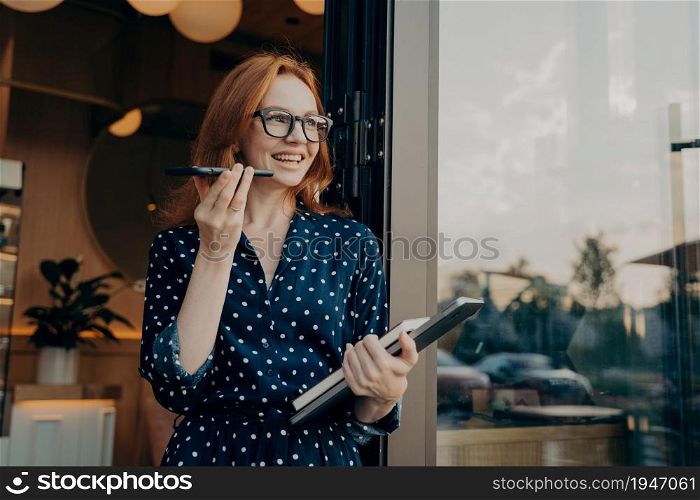 Happy cheerful ginger business woman freelancer with laptop holds mobile phone and talks on speakerphone with friend or client, uses virtual assistant or records audio message on smartphone outdoors. Happy ginger business woman freelancer with laptop holds mobile phone and talks on speakerphone