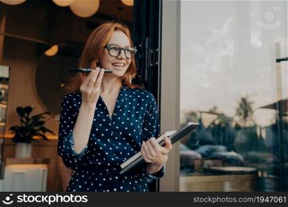 Happy cheerful ginger business woman freelancer with laptop holds mobile phone and talks on speakerphone with friend or client, uses virtual assistant or records audio message on smartphone outdoors. Happy ginger business woman freelancer with laptop holds mobile phone and talks on speakerphone