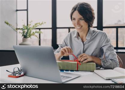 Happy cheerful Italian woman home office worker sits at desk unpacks newly received New Year gift from friends or grateful customers during Christmas Time, looking with broad smile at laptop screen. Happy cheerful Italian woman home office worker sits at desk unpacks newly received New Year gift