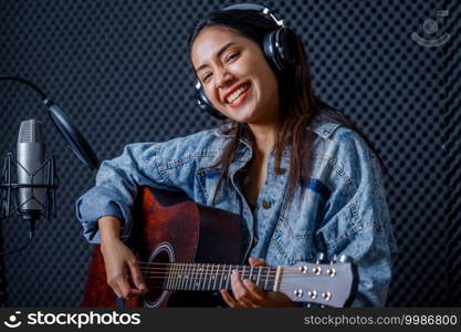Happy cheerful pretty smiling of portrait a young Asian woman vocalist Wearing Headphones with a Guitar recording a song front of microphone in a professional studio